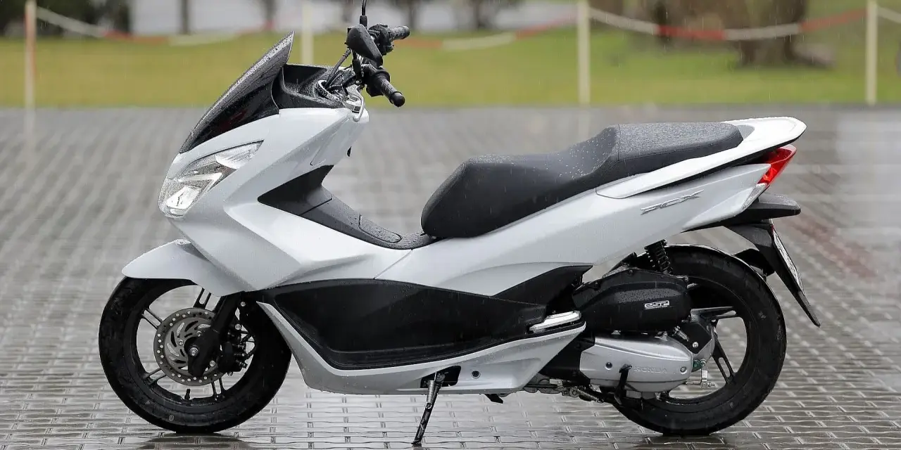 Biały skuter Honda PCX na mokrym bruku. Zastanawiasz się, od ilu lat można jeździć skuterem 50? Od 14 roku życia z kartą motorowerową.